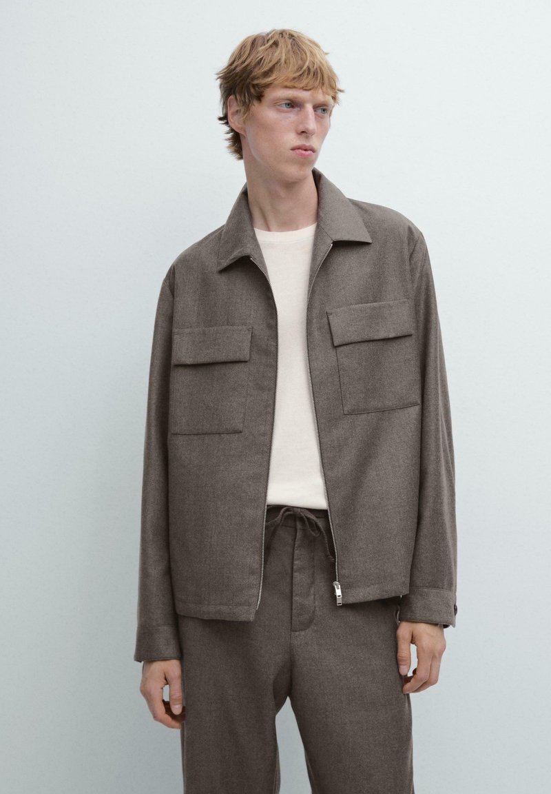 Giacca marrone con zip, realizzata in tessuto strutturato, con due grandi tasche sul petto, indossata sopra una camicia di colore chiaro, abbinata a pantaloni coordinati.