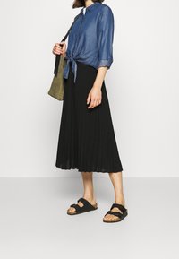 Denimskjorte med knyt foran, sort plisseret midi-nederdel og sorte sandaler med dobbelte stropper. En olivengrøn skuldertaske fuldender outfittet.