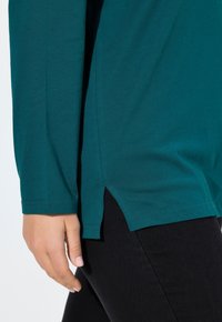 Teal langetermijnshirt van zachte stof met zijsplitten aan de zoom. Gedragen met zwarte slim-fit broek. Close-up van een hand die naast het shirt rust.