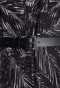 Ceinture en cuir noir avec boucle argentée attachée par-dessus une chemise à boutons à motif de feuilles de palmier noir et blanc.