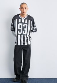 Mees, kellel on habeteta pea, seisab käed selja taga, kandes musta ja valge triibulist jersyt numbriga 93 ning lohvakate mustade pükstega, valge seina taustal.