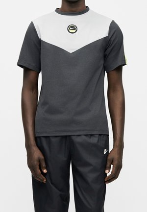 Mężczyzna noszący krótki rękaw koszulki Nike z białą górną częścią i ciemnoszarym dolnym pasem, w połączeniu z czarnymi spodniami Nike.