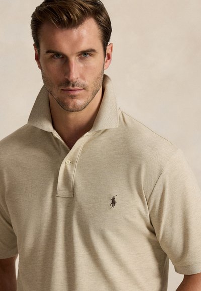 Mies, jolla on lyhyet ruskeat hiukset ja beige poolopaita, jossa on kohotettu kaulus ja kirjailtu polopelaajan logo.