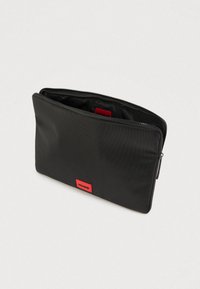 Etui dreptunghiular negru cu fermoar, cu un patch roșu cu logo-ul „HUGO” pe față, prezentat parțial deschis pe un fundal simplu deschis.