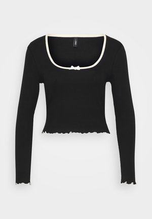 Must vahemütsiga must pikkade varrukatega topp, millel on kreemivärvi ääristus ja väike paeladetail. Omab lainelist tukka ja soonilist tekstuuri.