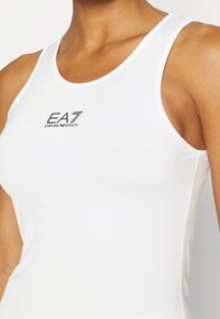 Närbild av en person som bär en vit ärmlös sporttopp med "EA7 Emporio Armani"-logotyp på bröstet.