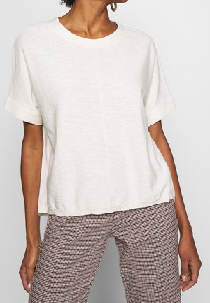 Woman wearing a loose-fitting, short-sleeve white top with a central seam, paired with red and white checkered pants.