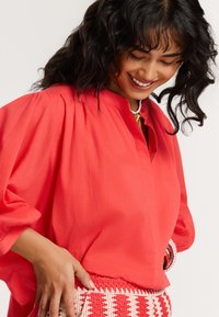Blusa leggera color corallo con scollo a V e maniche leggermente a sbuffo, abbinata a una gonna a fantasia con texture. Tessuto morbido con una vestibilità comoda.