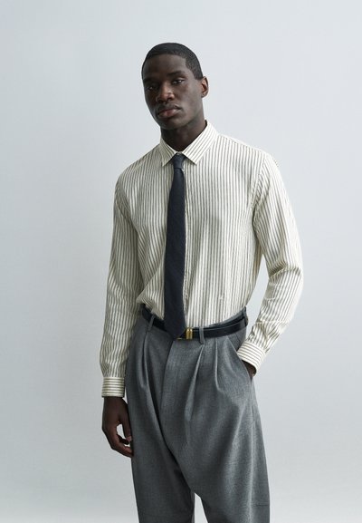 Homme portant une chemise crème à rayures, une cravate noire, un pantalon gris plissé et une ceinture noire, debout avec une main dans la poche devant un fond uni.