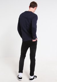 Uomo con jeans neri, maglione blu navy e sneakers Adidas bianco e nero, in piedi rivolto di spalle con una mano nella tasca posteriore.