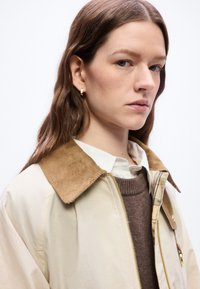 Γυναίκα με μακριά καστανά μαλλιά που φοράει μπεζ μπουφάν με καφέ γιακά από κοτλέ, καφέ πουλόβερ, λευκό πουκάμισο με γιακά και χρυσά σκουλαρίκια κρίκους.