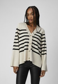 Beige strikket cardigan med sorte vandrette striber, V-hals, knaplukning foran og brede ærmer. Sammen med sorte tætsiddende bukser.