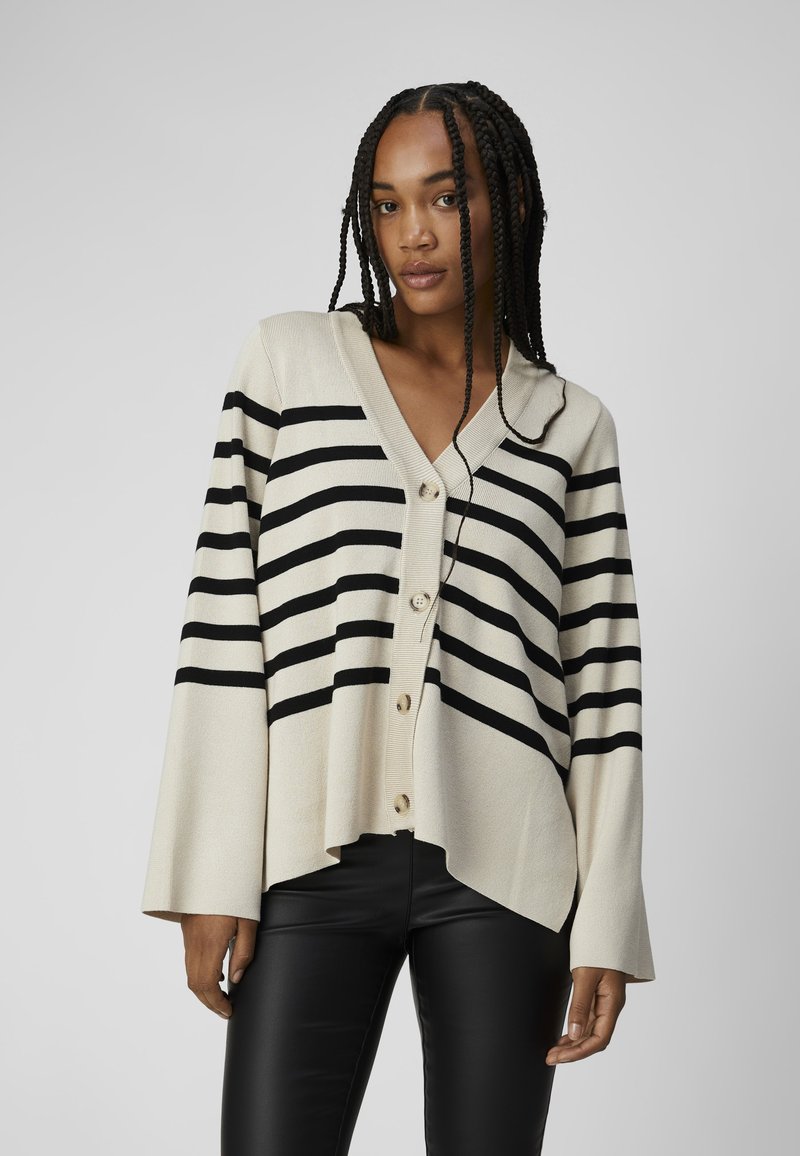 Beige strikket cardigan med sorte vandrette striber, V-hals, knaplukning foran og brede ærmer. Sammen med sorte tætsiddende bukser.
