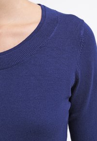 Mörkblå stickad tröja med vid halsringning och ribbad krage. Mjukt material och figurnära design som visar en slät tygfinish.