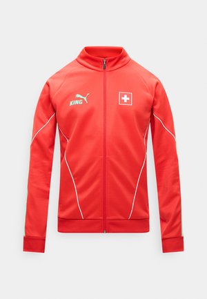 Crvena sportska jakna s cijelim patentnim zatvaračem, s logom Puma "KING" na lijevoj strani prsa i bijelim grbom križa na desnoj strani prsa, s bijelim detaljima obruba.