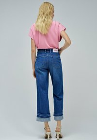 Camiseta rosa de manga corta, vaqueros azul de cintura alta con puños enrollados y sandalias de tacón beige. Marca notable en la cinturilla trasera.