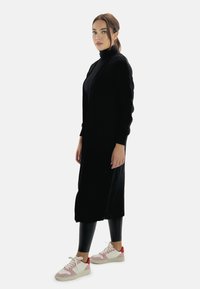 Cardigan nero a maglia con texture a coste, maniche lunghe, indossato sopra un dolcevita nero. Abbinato a leggings neri lucidi e sneakers con dettagli rosa.