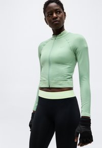 Ευλύγιστη γυναίκα με ζακέτα μακρυμάνικη σε μέντα, μαύρα κολάν με φαρδύ ανοιχτό πράσινο λάστιχο και μαύρα γάντια χωρίς δάχτυλα.