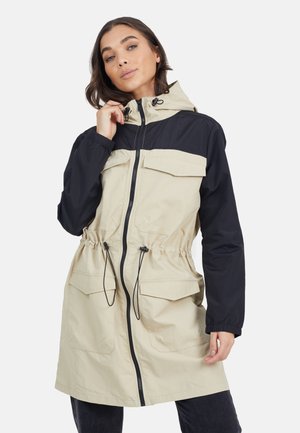 Femme portant une veste coupe-vent à capuche beige et noire avec fermeture éclair frontale, cordons de taille ajustables et poches à rabat, posant devant un fond uni.