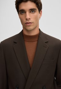 Bruine dubbelgeknoopte blazer met pieklapellen over een getextureerde bruine coltrui. Voorzien van een linkerborstzak en zwarte knopen.