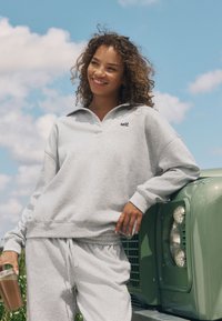 Helehall halli kraega ja logoga dressipluus, millele on sobitatud samas toonis dressipüksid. Modell hoiab selges klaasis jooki ja seisab rohelise sõiduki kõrval.