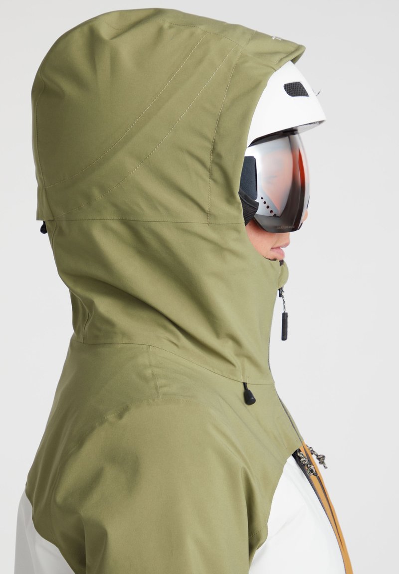 Skijacke mit grüner Kapuze, weißem Körper und braunen Akzenten. Verfügt über eine Reißverschluss-Tasche und besteht aus glattem, wasserabweisendem Material.