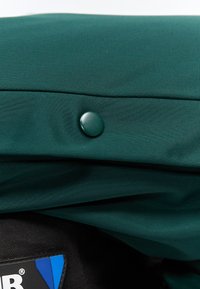 Sötétzöld anyag egyetlen kerek gombbal egy fekete anyagrész fölött, amely egy kék-fehér logófolt részletét ábrázolja.