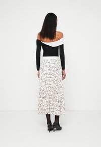 Fekete váll nélküli topp hosszú ujjakkal, fehér díszítésekkel és egy légies krémszínű szoknya fekete kézzel írt mintákkal. Fekete magassarkú cipő.