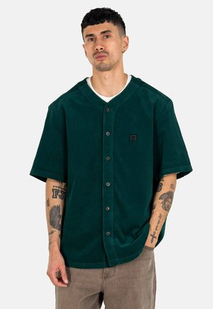 Dunkelgrünes Cordhemd mit kurzen Ärmeln, Knopfleiste, V-Ausschnitt und kleinem schwarzen Logo auf der Brust. Strukturierter Stoff und lässige Passform.