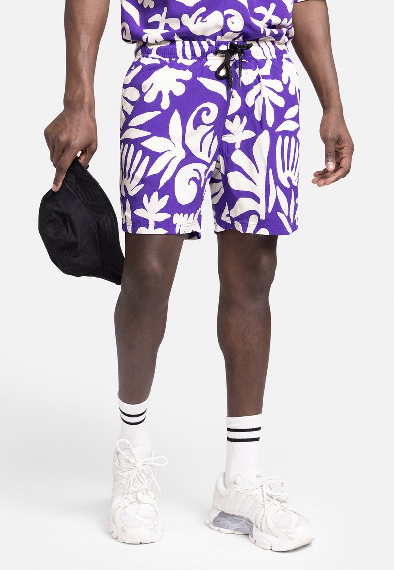 Homme portant un short rayé violet et blanc, des baskets blanches avec des chaussettes à rayures noires, tenant une casquette noire dans une main.