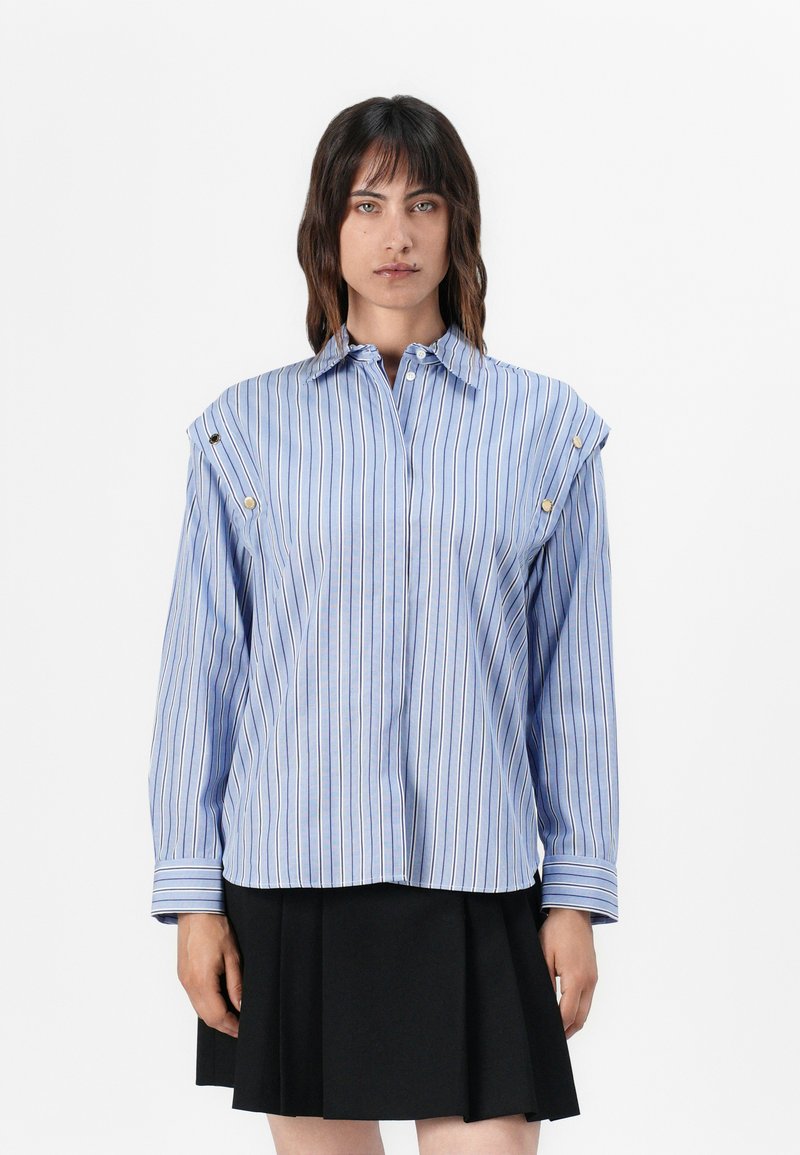 Vrouw in een blauw gestreepte blouse met lange mouwen en gouden knopen op de schouders, in een zwarte geplooide rok, staand voor een witte achtergrond.