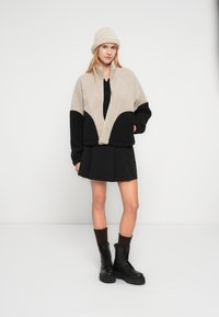 Νεαρή γυναίκα με μπεζ και μαύρο fleece μπουφάν, μαύρη φούστα, μπεζ σκουφάκι και μαύρα μποτάκια με κορδόνια και σκουρόχρωμες κάλτσες, στέκεται μπροστά σε λευκό τοίχο.