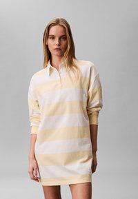 Femme portant une mini-robe à manches longues et col, avec de larges rayures horizontales beige et blanches, debout devant un fond gris uni.