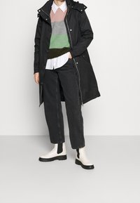 Cappotto nero lungo con cappuccio, indossato sopra un maglione a strisce multicolori lavorato a maglia, camicia bianca, jeans neri e stivaletti bianchi con accenti neri.