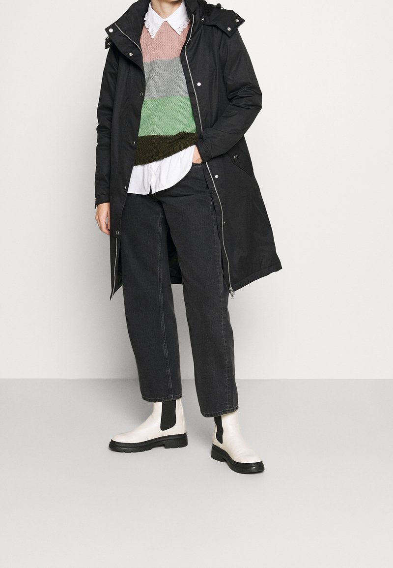 Cappotto nero lungo con cappuccio, indossato sopra un maglione a strisce multicolori lavorato a maglia, camicia bianca, jeans neri e stivaletti bianchi con accenti neri.
