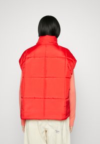 Röd fluffig väst med hög krage, överdimensionerade axlar och quiltat mönster. Slät textur, avklippta ärmar, bärs över en ljusrosa topp.