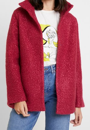 Personne portant une veste rouge texturée ouverte sur un t-shirt blanc avec un motif abstrait de visage en ligne et un jean bleu.