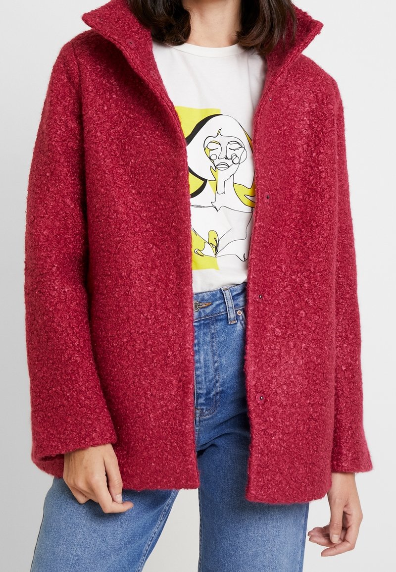 Personne portant une veste rouge texturée ouverte sur un t-shirt blanc avec un motif abstrait de visage en ligne et un jean bleu.