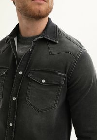 Zwarte denimjas met twee borstzakken, zilveren drukknoopjes, een western-stijl yoke en een versleten afwerking, gedragen over een grijze trui.