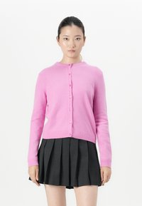 Jongeman met donker haar in een roze gebuttonde cardigan en een zwarte plissé rok staat tegen een witte achtergrond, met het gezicht naar voren.