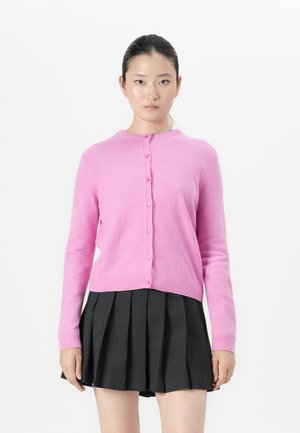 Jeune femme aux cheveux bruns, portant un cardigan rose à boutons et une jupe plissée noire, se tient face à l'objectif sur fond blanc.