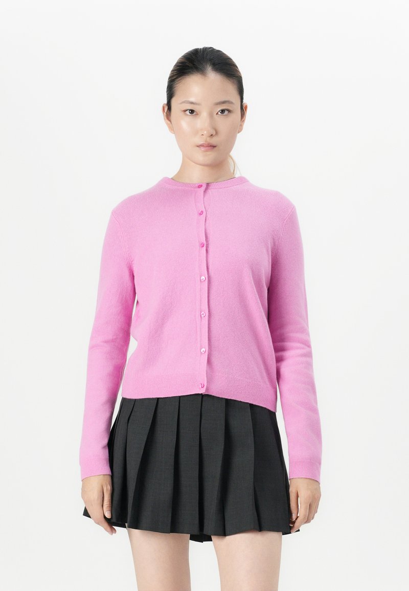 Jongeman met donker haar in een roze gebuttonde cardigan en een zwarte plissé rok staat tegen een witte achtergrond, met het gezicht naar voren.