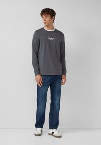 Graues Langarmshirt aus Baumwolle mit Rundhalsausschnitt und Logo auf der Vorderseite. Kombiniert mit blauen Jeans und weißen Sneakern mit schwarzen Akzenten.