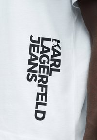 Λευκό ένδυμα με μαύρο κάθετο κείμενο «KARL LAGERFELD JEANS» που φοριέται από άτομο με σκούρο χρώμα δέρματος, ορατό στη δεξιά πλευρά.