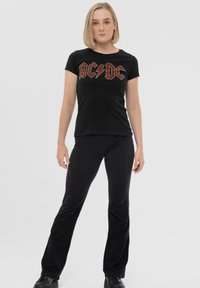 Black t-shirt with red and yellow AC/DC logo, short sleeves, and crew neck, paired with black pants and black shoes, standing on a light background.