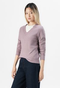 Jovem mulher com cabelo loiro longo, vestindo um suéter V, roxo claro, sobre uma blusa branca e jeans azul escuro, de pé contra um fundo branco.
