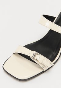 Sandal de charol crema con dos correas ajustables y punta cuadrada. Plantilla texturizada negra y tacón negro delgado. Detalle de hebilla de metal plateado.