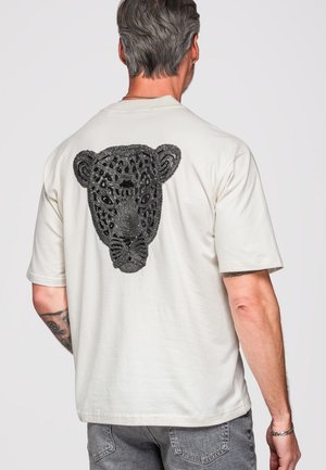 Uomo che indossa una maglietta bianca con un volto di leopardo nero ricamato dettagliatamente sulla schiena, in piedi davanti a uno sfondo semplice.