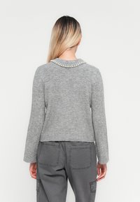 Žena s dlhými blond vlasmi, otočená chrbtom, oblečená v šedom pletenom svetri s perlovými detailmi na golieri a tmavosivých cargo nohaviciach.