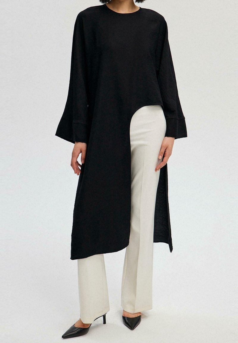 Haut noir à manches longues avec un ourlet incurvé et une fente latérale, associé à un pantalon blanc à jambes droites et des escarpins noirs à bouts pointus. Texture lisse.